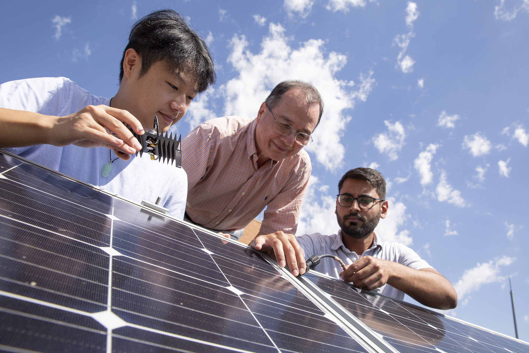 Three men lean over a large solar panel, one holding a part and one adjusting a cable, with a beautiful blue sky overhead.