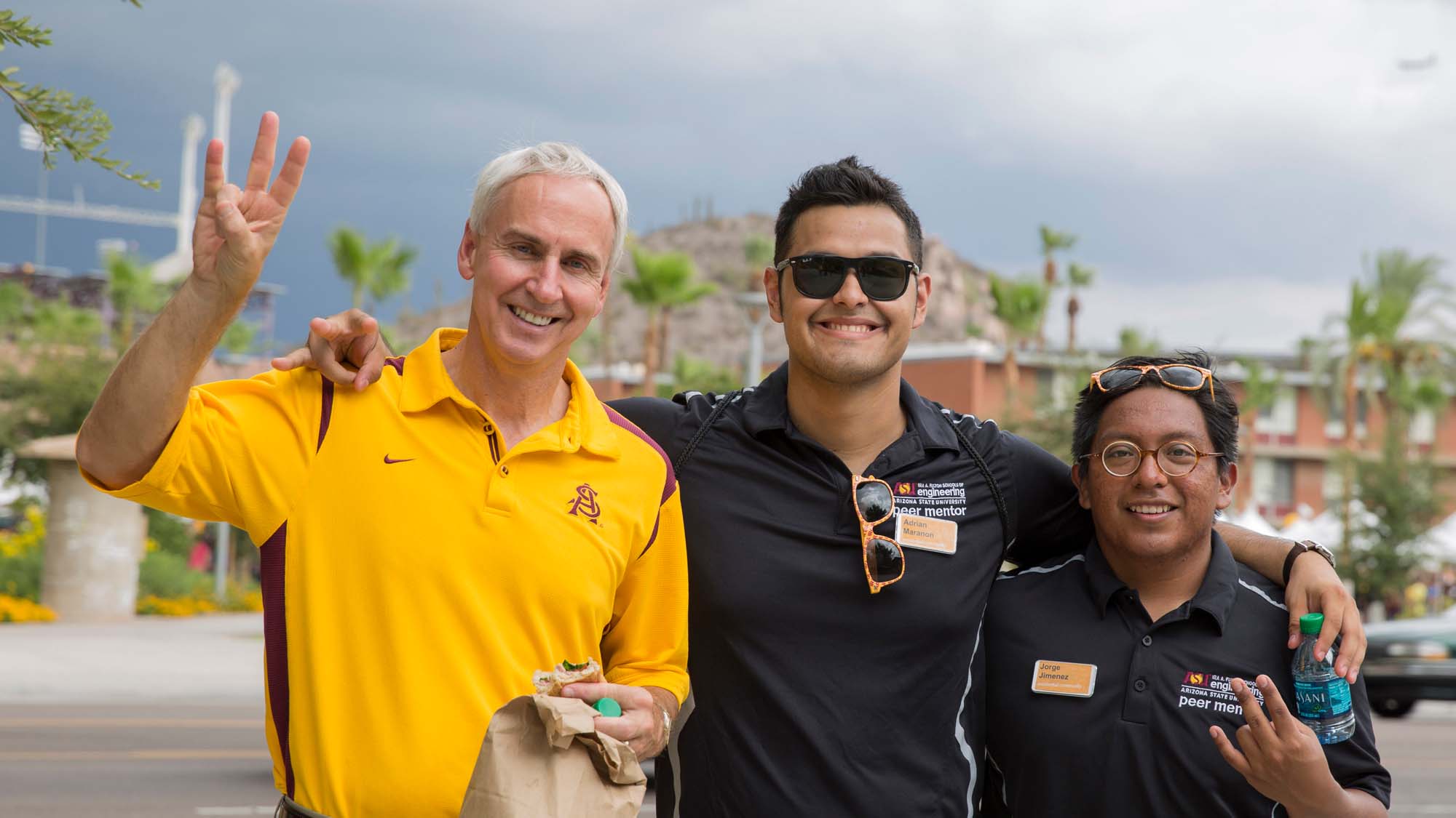 Three cheerful individuals in polo shirts pose at a university event, with tropical foliage and a cloudy sky in the background.