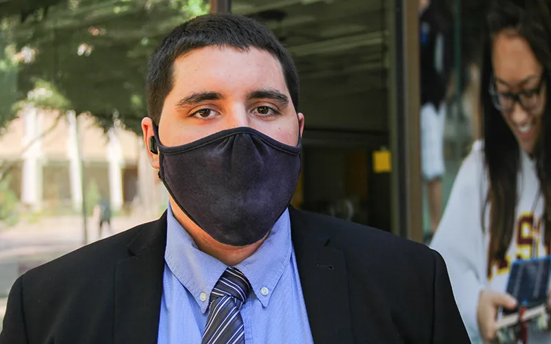 A man in a dark suit and blue shirt looks at the camera. He is wearing a black face mask.