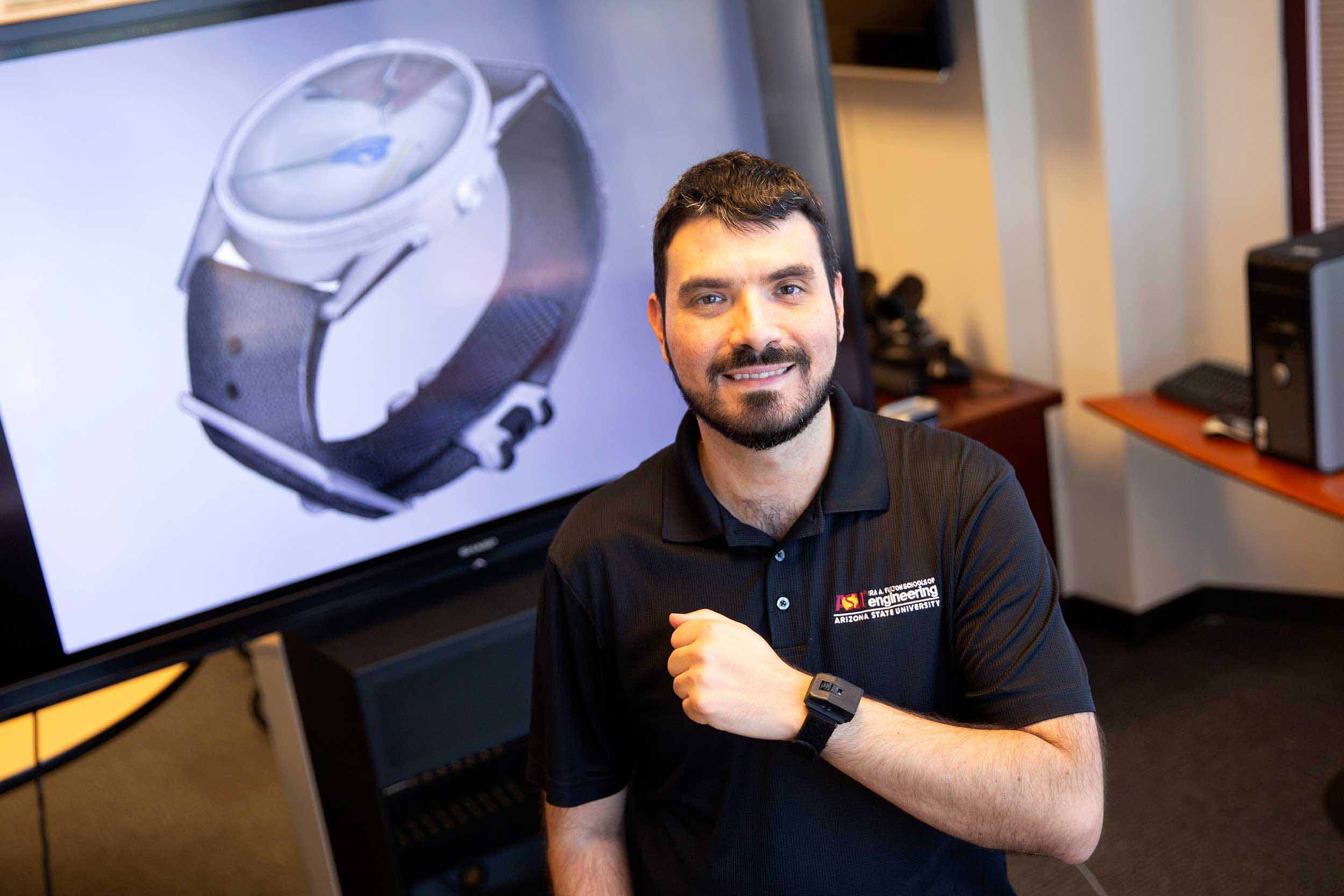 Troy McDaniel stands looking at the camera in his lab, displaying his wrist device that showcases his technology