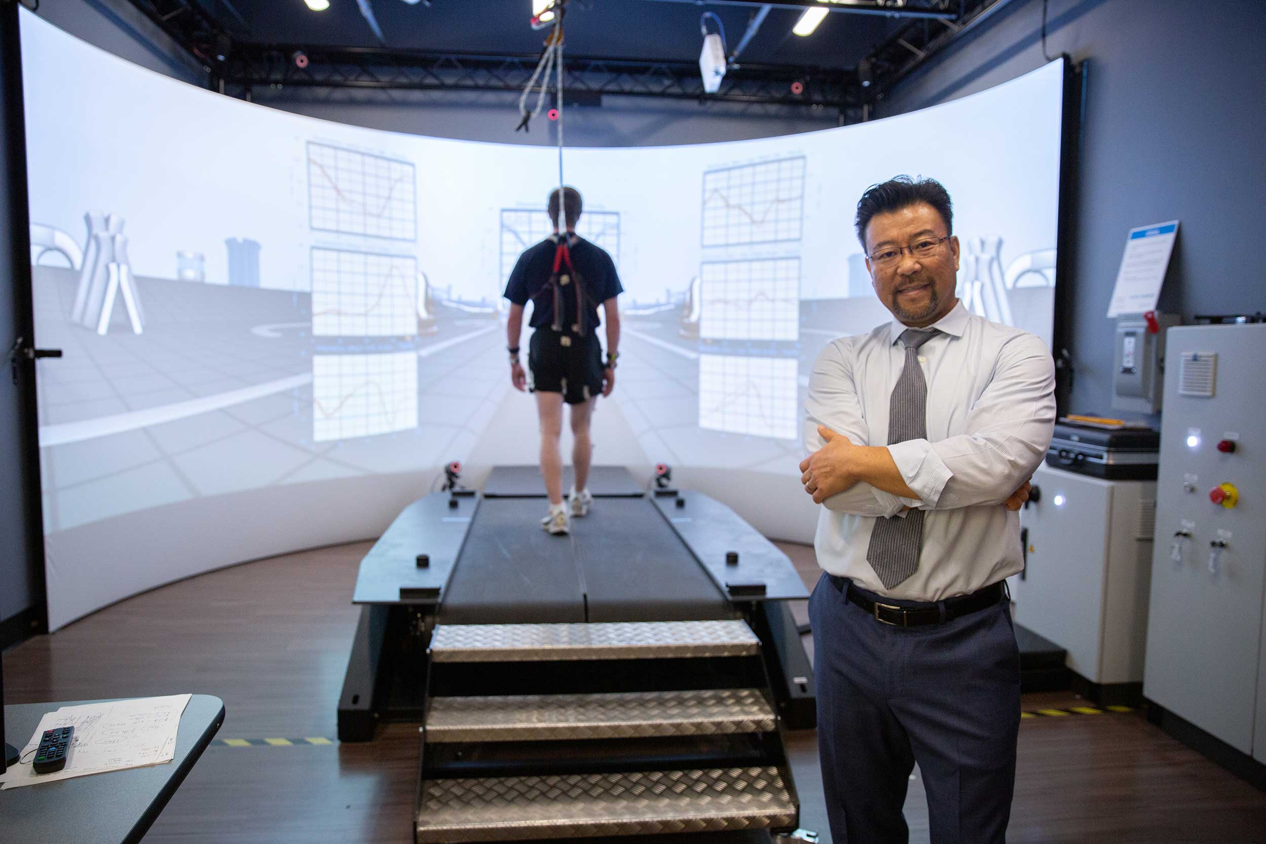 Thurman Lockhart stands in his mobility lab, while a person walks on a specialized treadmill connected to a digital apparatus walks in the background