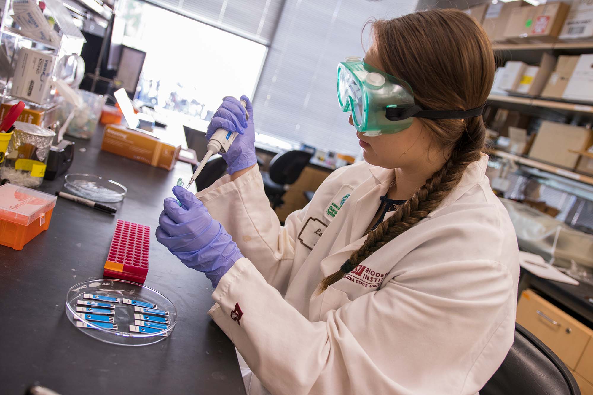 A female student wearing a lab coat and protective gear working on her research project at ASU research facilities.
