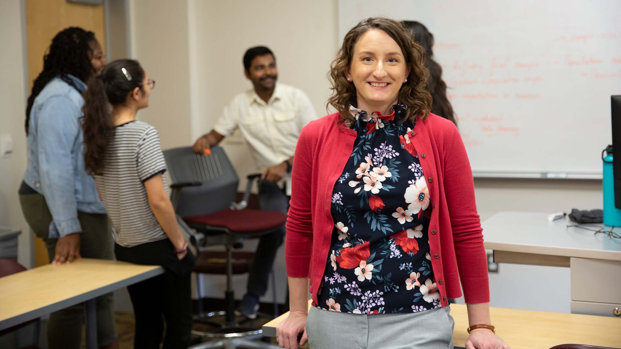 Portrait of Samantha Brunhaver in her classroom with students chatting behind her near the white board.