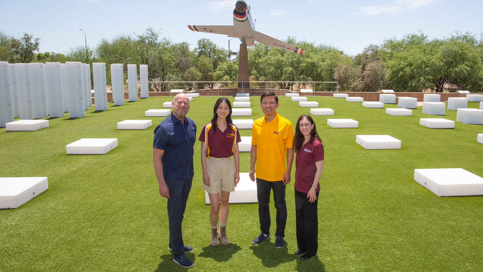 Four researchers stand together on bright green turf under the bright Arizona Sun among a field of white boxes that are arranged at even intervals on the turf.