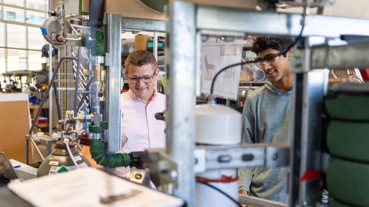 Two people stand in a laboratory among various industrial and scientific equipment, looking at a monitor and smiling.