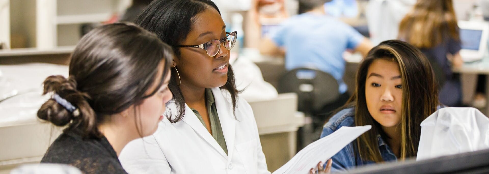 Three students in a lab setting review a document together, with one student in a white lab coat explaining to the others.